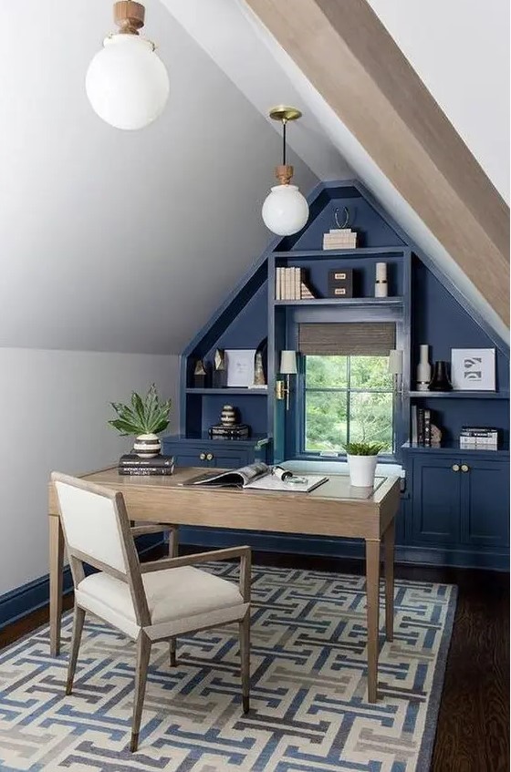 An elegant farmhouse attic home office with a blue accent wall with built in storage, a stained desk and a neutral chair, pendant lamps