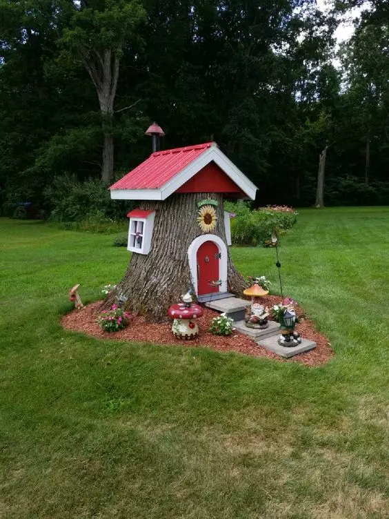 a small tree stump elf house with a red roof and windows, a red door and some blooms and gnome figurines is amazing