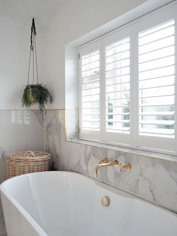 a modern neutral bathroom clad with marble tiles, an oval tub, gold fixtures and a window with shutters plus potted greenery