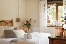 a neutral and cozy Mediterranean bedroom with stained wooden beams, a bed with neutral bedding, a stained dresser, pendant lamps, candle lanterns and a jute pouf