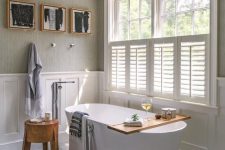 a welcoming neutral bathroom with grey and creamy paneled walls, a window partly covered with shutters, an oval tub, a wooden stool