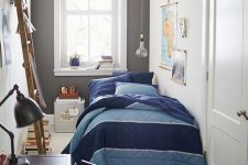 08 a narrow kid’s room with a bed with blue and navy bedding, a dark-stained desk and a chair, a black rug and a ladder, some artwork