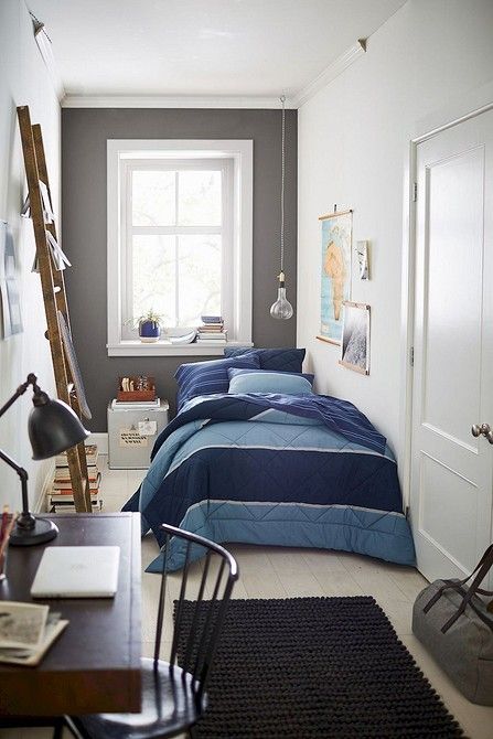 A narrow kid's room with a bed with blue and navy bedding, a dark stained desk and a chair, a black rug and a ladder, some artwork