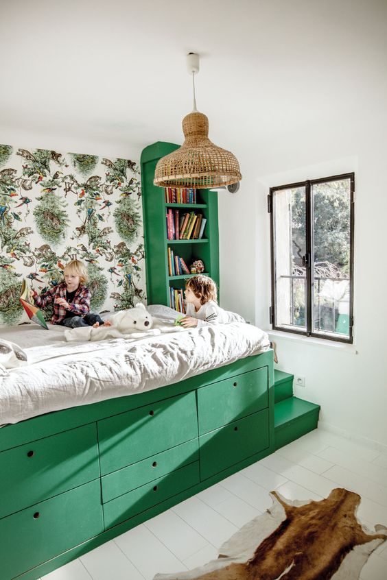 a small and bright kids' room with a green storage bed and a built-in bookshelf, a printed wallpaper wall and a woven pendant lamp