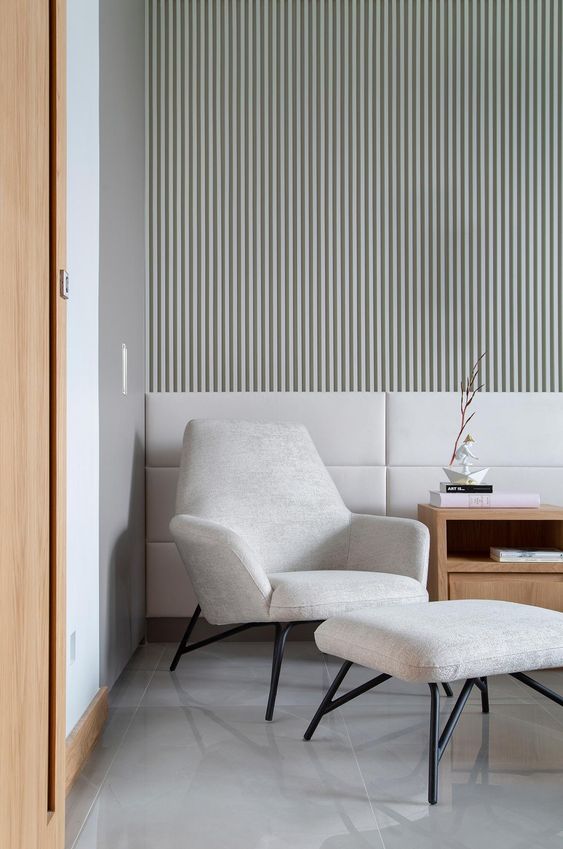 A contemporary space with a whitewashed wood slat accent wall, upholstered panels, a neutral chair with a footrest and a light stained side table
