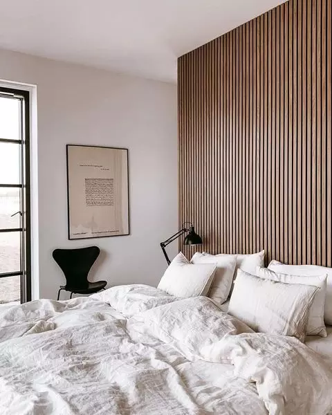 a neutral bedroom with a wood slat accent wall, a bed with neutral bedding, a black chair, a black table lamp and a print on the wall