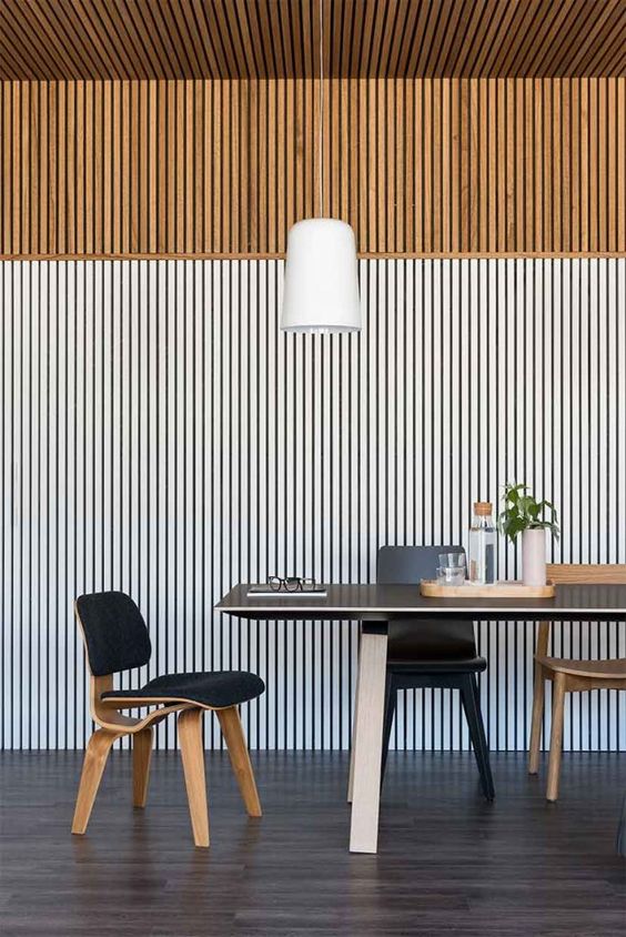 A mid century modern dining room with a stained and whitewashed wood slat wall, a black table and black and stained chairs, a pendant lamp