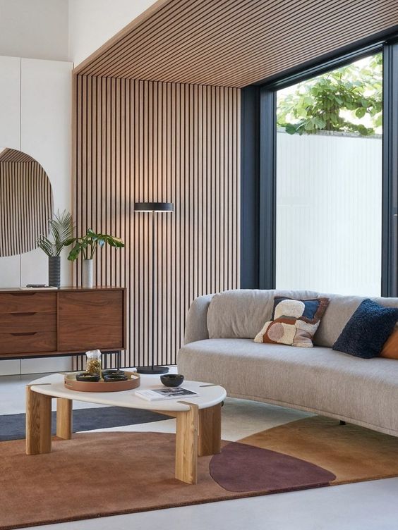 An elegant mid century modern living room with a wood slat accent wall and ceiling, a curved sofa, a printed rug, a stained credenza and a coffee tabl