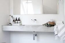 a Scandinavian bathroom done in white, with a floating concrete sink, a mirror cabinet, a wooden stool and a simple metal trash can