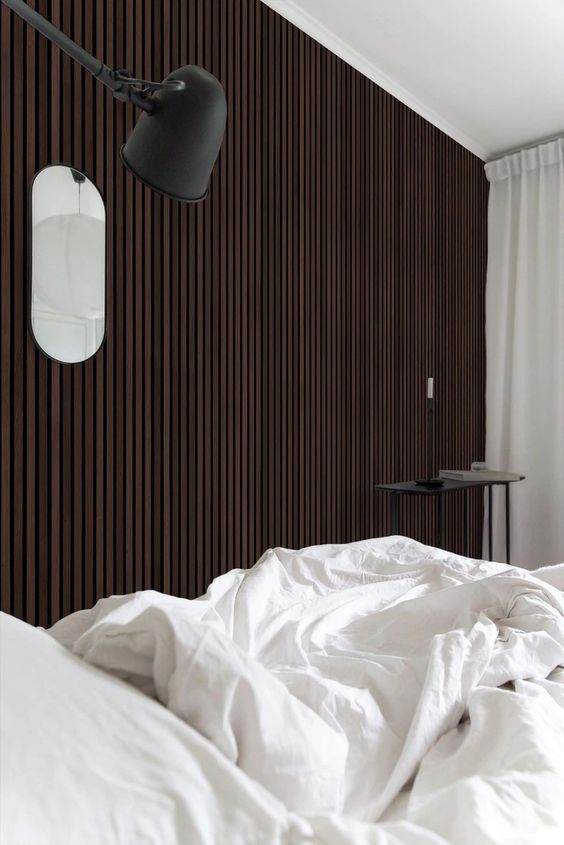 A catchy bedroom with a dark stained wood slat accent wall, a bed with neutral bedding, a black coffee table and a black sconce