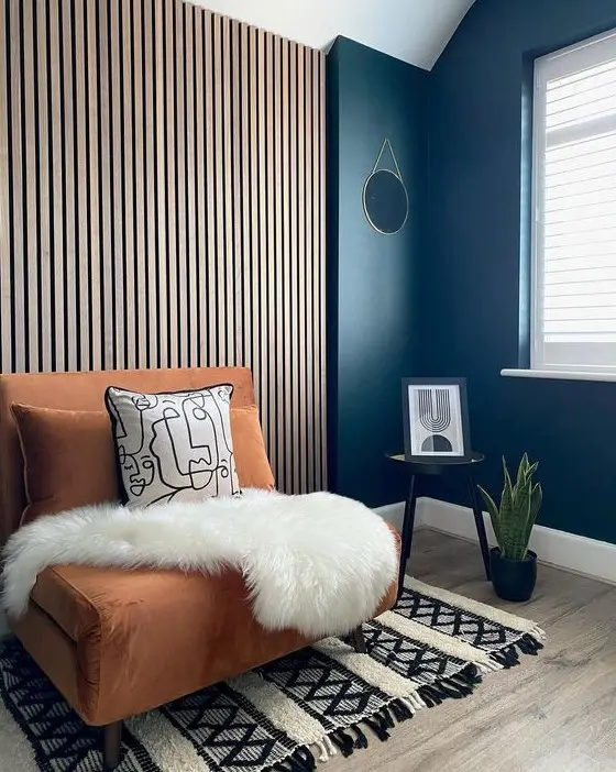 A chic mid century modern nook with a wood slat accent wall, a rust colored chair with pillows, a printed rug, a side table and a potted plant