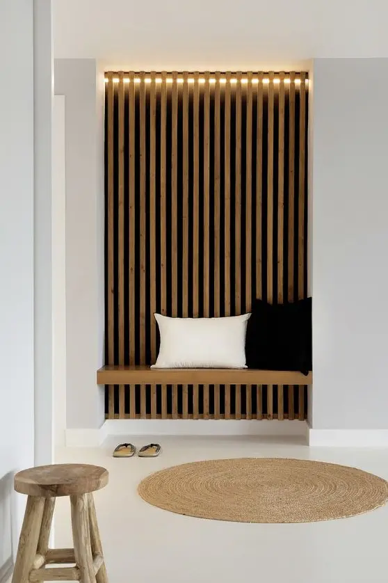 A laconic Japandi entryway with a wood slat accent wall and lights, a built in daybed and a wooden stool and jute rug