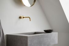 a minimalist attic bathroom featuring a floating grey stone sink, brass fixtures and a round mirror plus lots of natural light