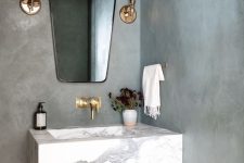 a modern bathroom with cocnrete walls, a small yet chic marble slab floating sink, brass fixtures and a catchy mirror