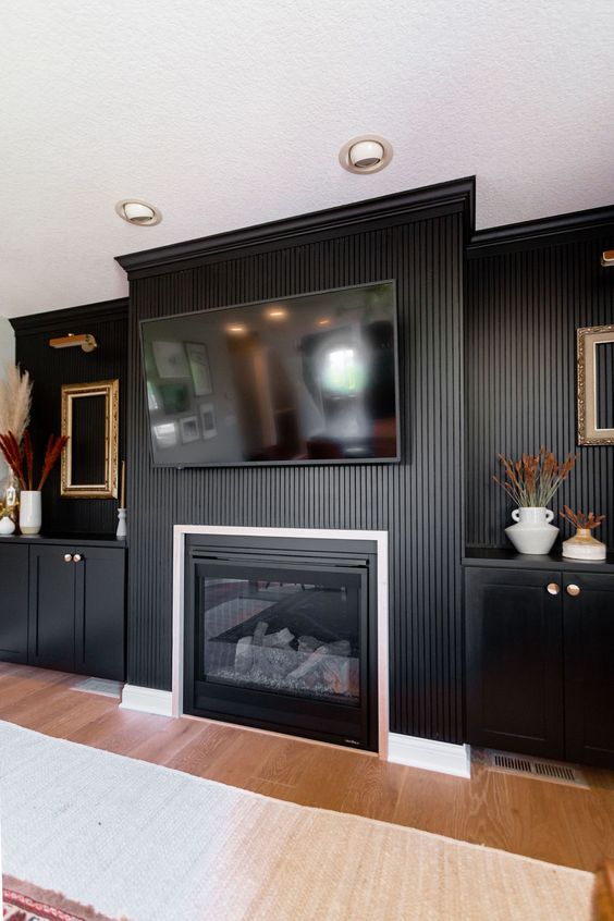 A refined living room with black walls and a wood slat accent one, a built in fireplace and black cabinets, gold touches
