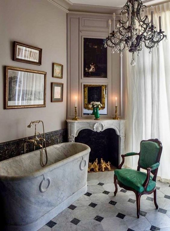 A refined vintage bathroom with a stone bathtub, a non working fireplace, a green chair, a refined crystal chandelier and a gallery wall