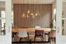 a stylish dining room with a wood slat wall, a stained table, mismatching chairs, a cluster of pendant lamps and potted plants