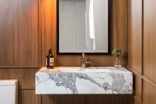an elegant powder room with wood panels on the walls, a floating white marble sink, a catchy chevron marble floor and a black sconce