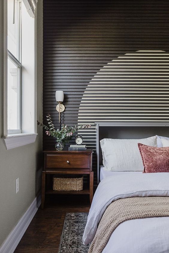 An eye catching wood slat accent wall with a painted circle, a grey bed with bedding, a stained nightstand and some printed rugs