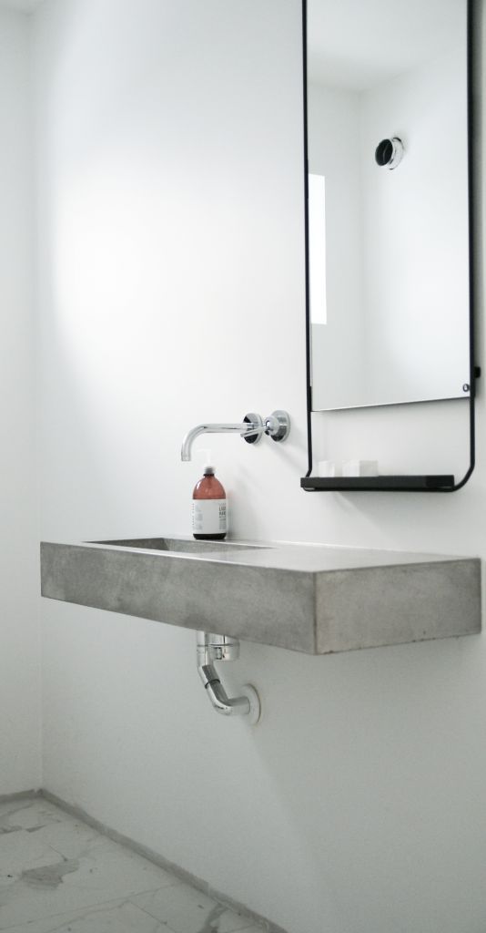 An ultra minimalist bathroom with white walls, a concrete floating sink, a catchy mirror with an additional small shelf