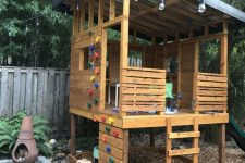 14 a kids’ playhouse of wood, with a roof and some furniture, a colorful cimbing wall, a ladder and some planters around is a cool space
