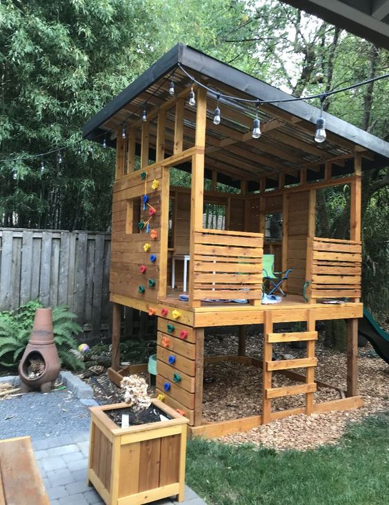 a kids' playhouse of wood, with a roof and some furniture, a colorful cimbing wall, a ladder and some planters around is a cool space