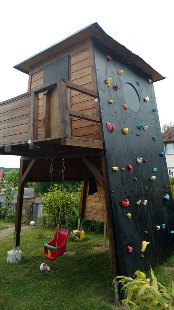 a kids' playhouse with a swing, a room on top and a black climbing and a round window is a lovely idea for a garden