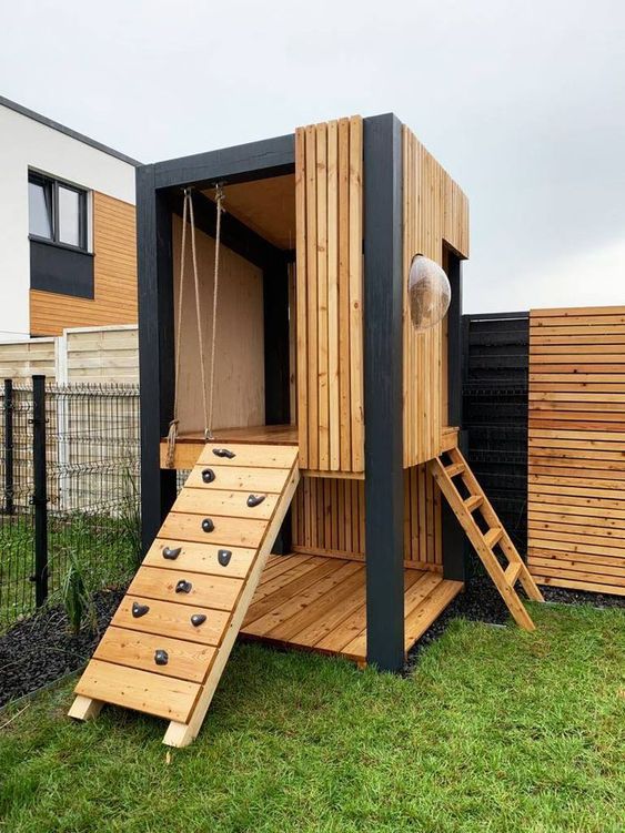 a modern light-stained kids' house with a ladder and a climbing wall and a sphertic window is a lovely idea