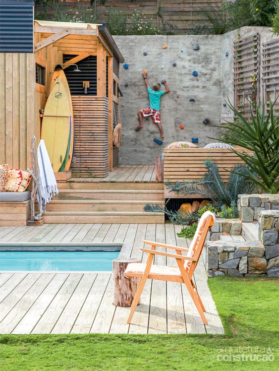 A modern outdoor space with a small pool, a wooden deck, built in and usual furniture of wood, a small outdoor shower and a climbing wall for kids