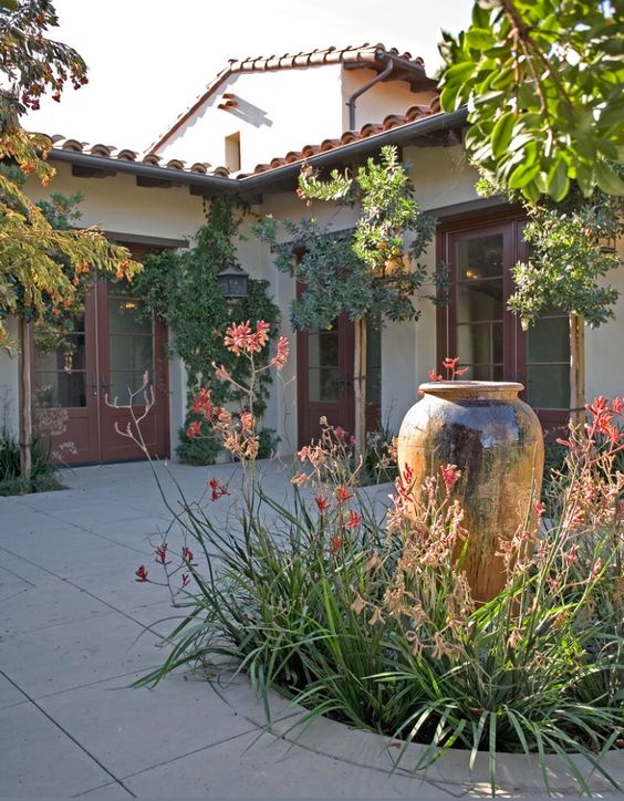 a beautiful front yard with stone tiles on the floor, an amphora-shaped fountain surrounded with greenery and blooms