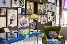 a bright and catchy home office with brown walls, a bold blue desk, a green chair with pillows, a colorful gallery wall and a large mirror