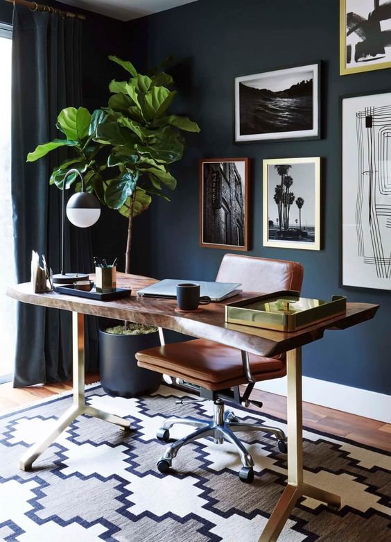 A chic mid century modern home office with navy walls and curtains, a living edge desk, a leather chair, a catchy gallery wall and a potted tree