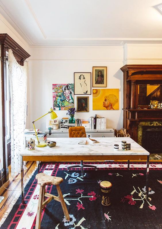 A colorful home office with a marble desk, a leather chair and a wooden stool, a built in fireplace, a credenza, a colorful gallery wall