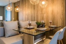 a contemporary dining nook with a mirror wall, a built-in and lit up upholstered taupe bench, a wood and glass table and white chairs
