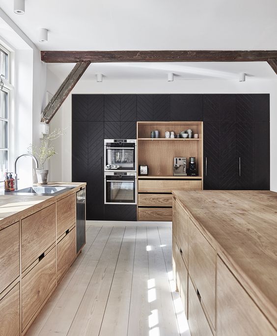 a contemporary kitchen with sleek light-stained cabinets, a whitewashed floor, a large black storage unit with built-ins and a wooden beams