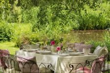 a delightful Provence outdoor space with a dining set of metal, with lots of cushions and pillows, bright blooms and greeneyr over it