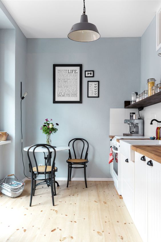a dusty blue kitchen with white cabinets, an open shelf, a round table, black chairs and a pendant lamp, a mini gallery wall