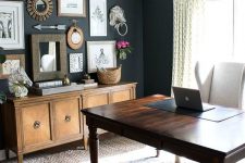a lovely vintage home office wiht black walls, a vintage dark-stained desk, a creamy chair, a credenza and a rustic gallery wall