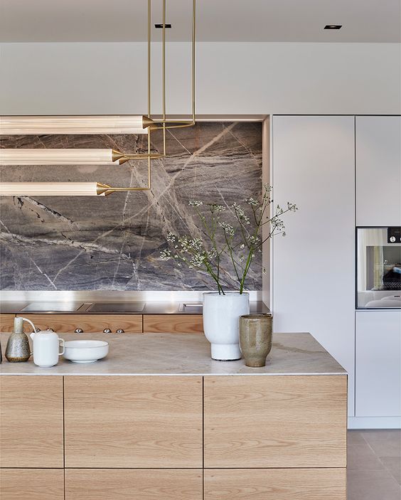 A luxurious minimalist kitchen with sleek light stained cabinets, a marble backsplash, built in appliances and a chic chandelier