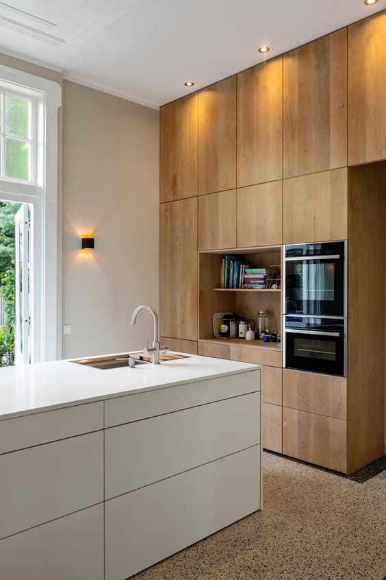 A minimalist kitchen with light stained no hardware cabinets and a white sleek kitchen island, built in appliances and lots of light