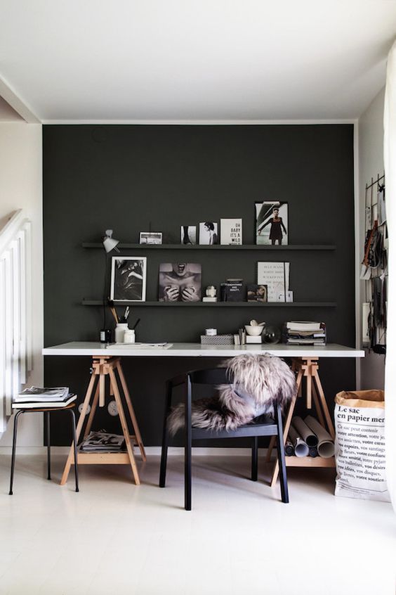 a moody home office with a black accent wall, a trestle deck, a black chair and a stool, a ledge gallery wall and a grid memo board