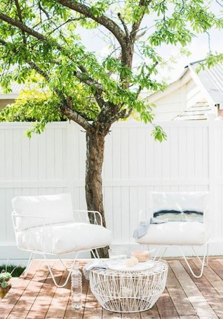 a small and cozy modern outdoor space with a wooden deck, white metal furniture with upholstery and a large living tree