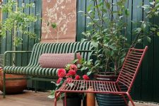 a stylish and bright terrace with a tall fence in green, a green metal sofa and a red chair, some potted plants and greenery