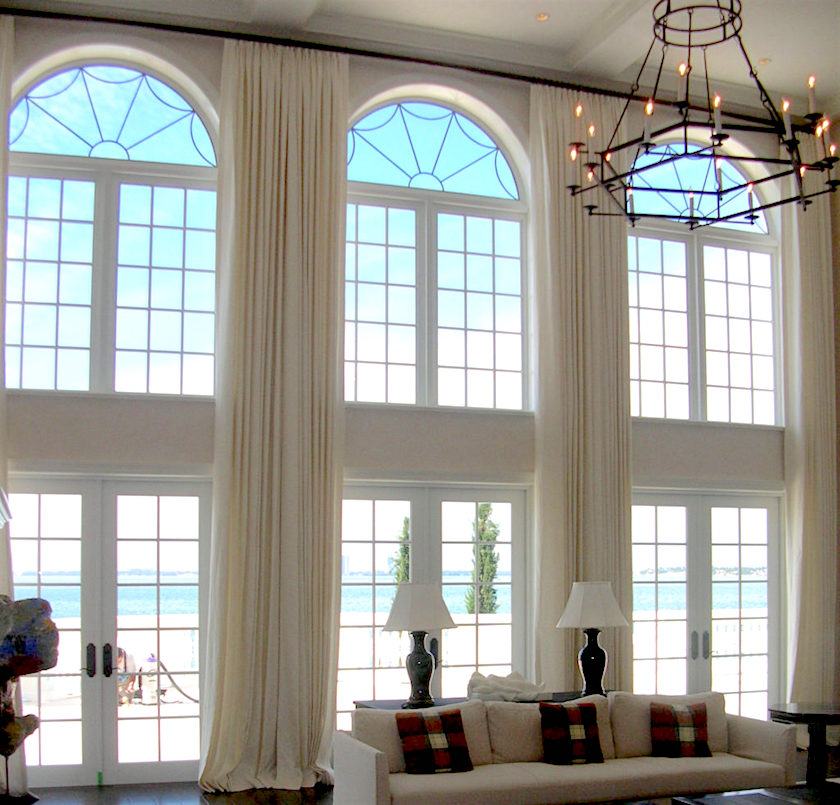 A modern living room with tan furniture, double height ceiling and windows with arched tops done with matching neutral drapes that cover everything