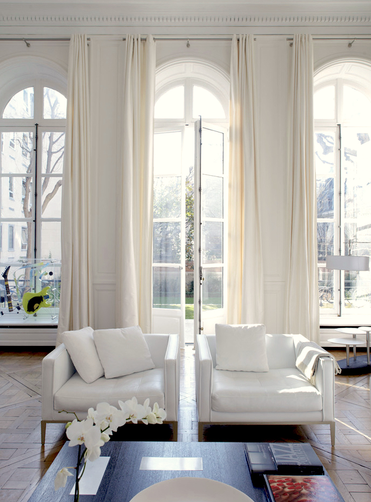 a sophisticated white living room with high ceilings, arched French windows, each of them done with creamy drapes that match