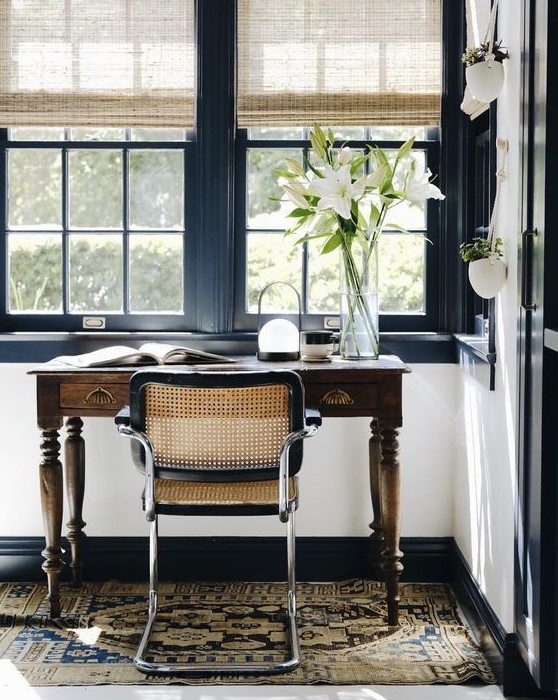 a beautiful home office with navy framed windows, a vintage stained desk, a rattan chair and shades plus a boho rug