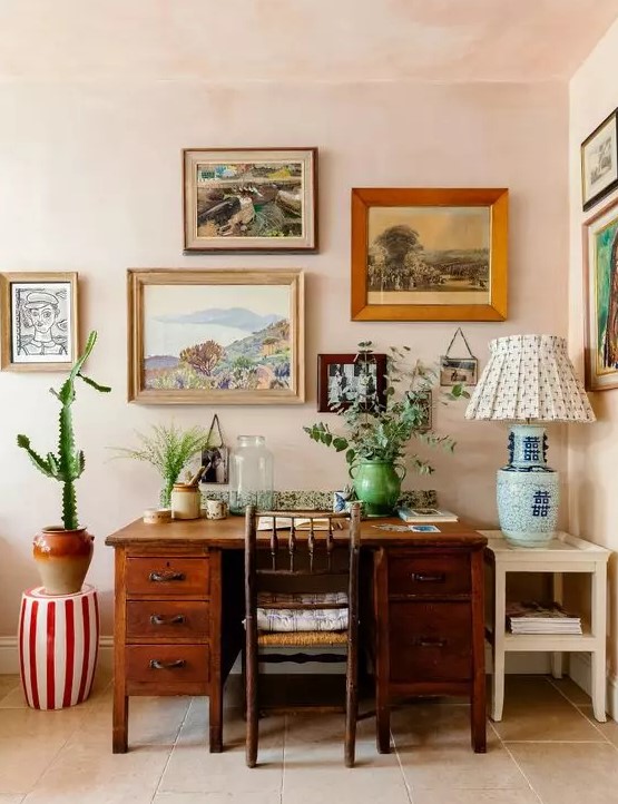 A bright mid century modern home office with blush walls, a stained desk and a chair, a colorful gallery wall and greenery and a cactus