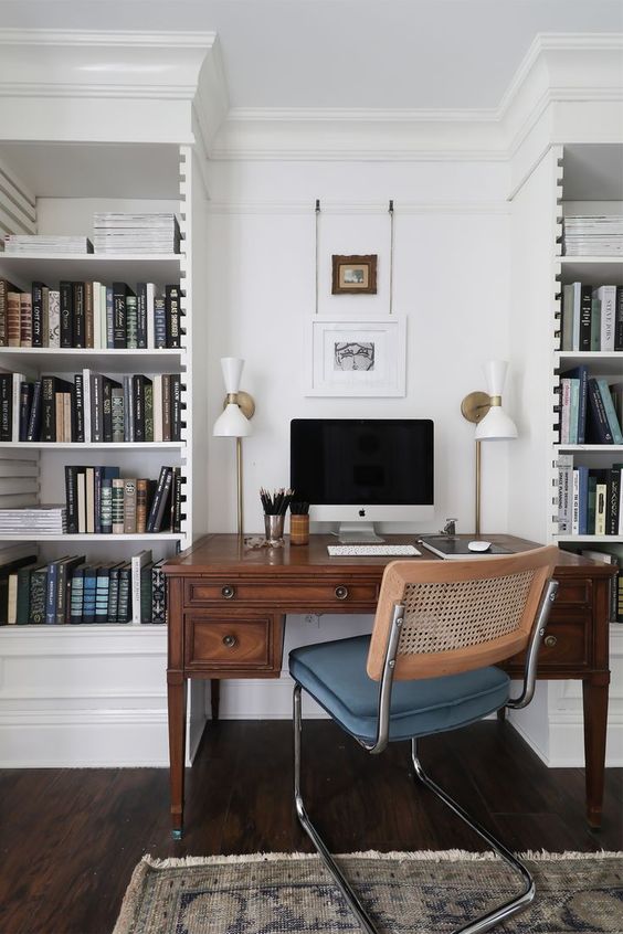 A chic home office with white built in bookcases, a stained vintage desk, a modern chair and some art and chic lamps is a cool space