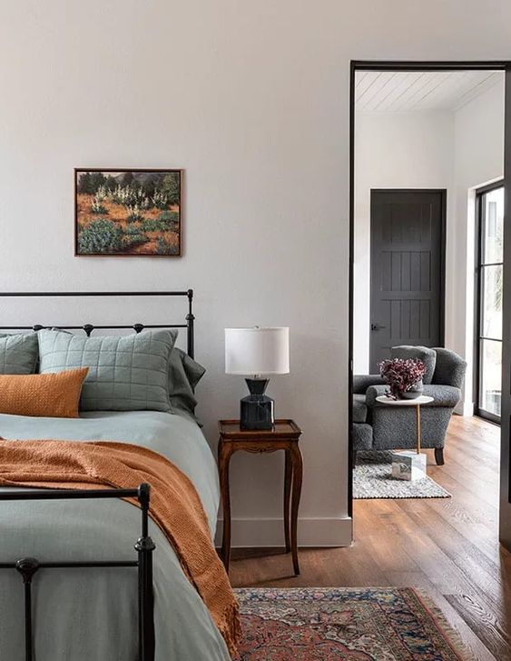 A modern bedroom with a black forged bed with muted green bedding, a dark stained vintage nightstand and a table lamp