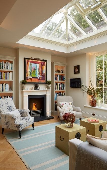 A modern neutral living room with a glass roof, a fireplace, built in bookshelves, a neutral sofa, yellow chests and vintage chairs